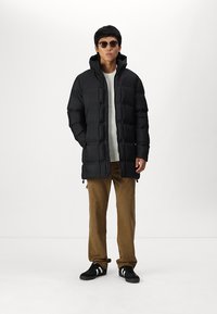 Schwarze Daunenjacke mit hohem Kragen, gesteppter Optik und zwei Seitentaschen. Getragen über einem weißen Pullover und kombiniert mit braunen Cordhosen.
