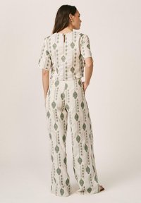 Femme portant une tenue blanche deux pièces avec des motifs de feuilles vertes, debout avec une main dans la poche, tournée de dos sur un fond uni.