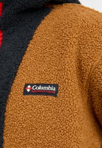 Pruun ja must karusnahkline jakk, millel on tekstureeritud, koheline pind. Esiküljel on Columbia Sportswear Company logo silt.