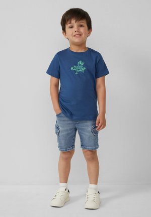 Granatowa bawełniana koszulka z grafiką zielonego dinozaura, zestawiona z dzinsowymi szortami i białymi sneakersami. Prosty design i luźny krój.