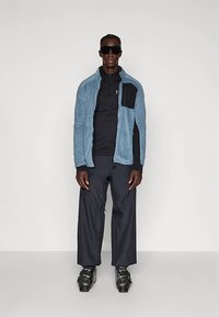 Man staat met zwarte zonnebril, blauwe ritsjack, zwarte broek en zwarte skischosten tegen een eenvoudige lichte achtergrond.