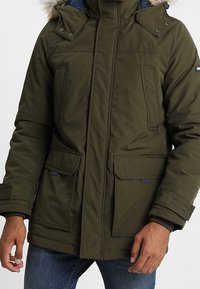 Olivgrön parkasjacka med huva i fuskpäls, tryckknappdetaljer, två frontfickor och dragkedjestängning. Texturerad tyg med ett lager på lager-design.