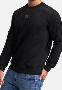 Mand iført sort sweatshirt med teksten "WEAR THE CODE" og blå jeans, hånd i lommen, mod hvid baggrund.