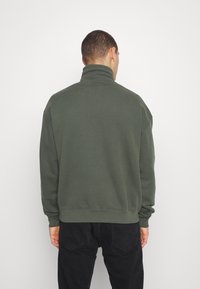 Sudadera de cuello alto en verde oscuro, fabricada con material suave, que presenta un ajuste relajado, puños acanalados y un dobladillo recto. Sin marcas visibles.