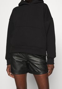 Czarna bluza z kapturem z przednią kieszenią i ściągaczem u dołu, zestawiona z błyszczącymi czarnymi skórzanymi szortami z podwiniętymi mankietami.