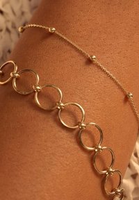 Bracelet en chaîne dorée comprenant des maillons circulaires entrelacés et une chaîne délicate avec de petites perles, posé sur une peau bronzée.