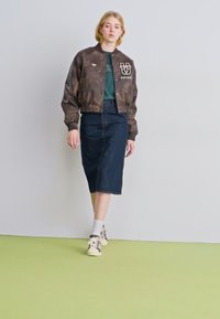 Casaco bomber castanho com emblemas, t-shirt verde, saia midi de ganga, meias brancas e sapatilhas; de pé num chão verde claro.