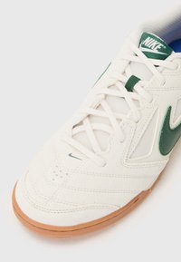 Hvit atletisk sko med grønne detaljer, mesh-paneler og strukturert semsket skinn. Har en brun gummisåle og signatur Nike-logo.