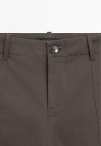 Brune bomuldsbukser med flad front, knaplukning og bæltestropper, med subtil diagonal tekstur og kontrasterende syninger.