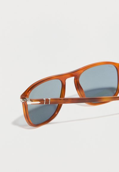 Smeđe prozirne sunčane naočale s tamnosivim pravokutnim staklima i detaljima srebrnih metalnih šarki, prikazane pod kutom na bijeloj pozadini.