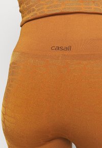 Pomarańczowe legginsy w wzór, wykonane z elastycznego materiału o metalicznym połysku, z gładkim pasem w talii, na którym znajduje się nadrukowana nazwa marki "casall".