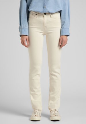 Personne portant un jean droit couleur crème, une chemise bleu clair boutonnée et des baskets crème à lacets, debout devant un fond uni.