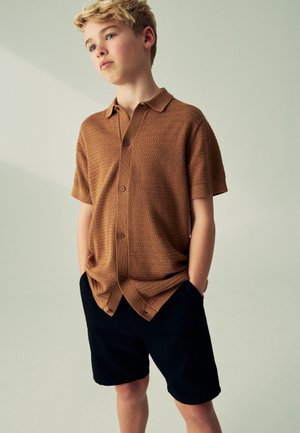 Blonder Junge trägt ein braunes kurzärmeliges Hemd mit Knöpfen und schwarze Shorts, steht mit den Händen in den Taschen und schaut leicht nach oben und zur Seite.