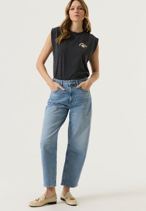 Femme debout les mains dans les poches, portant un haut graphique noir sans manches, un jean taille haute bleu clair et des mocassins beiges à pampilles.