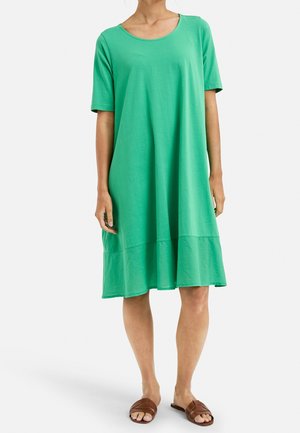 Femme portant une robe verte ample, longueur genou, avec des manches courtes et des sandales marron sans bride, debout devant un fond uni.