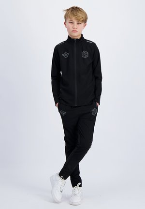 Blond kind staat met handen in de zakken, draagt een zwart trainingspak met logo's en witte sneakers tegen een effen lichte achtergrond.