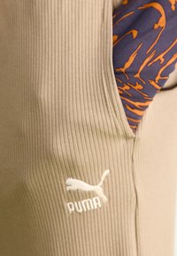 Stof ribbed bej cu un logo Puma în colțul din stânga jos; un articol bleumarin cu un model portocaliu se întrevede din lateral.