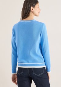 Pull bleu clair en matière tricotée douce avec un col rond. Les poignets et l'ourlet côtelés présentent des rayures blanches et bleu clair. Jean en denim foncé visible en dessous.