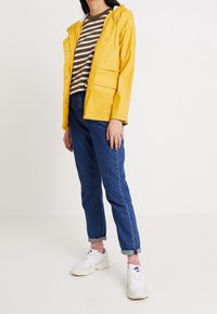 Veste imperméable jaune avec capuche, associée à une chemise rayée marron et blanche, un jean bleu et des baskets blanches. Design simple et poches fonctionnelles.
