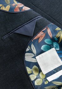 Tessuto scuro con una texture, accentato da motivi floreali in blu, verde e pesca, con una toppa bianca e bordi lisci color navy.