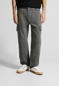 Šedé cargo kalhoty vyrobené z bavlny s volným střihem, které mají více bočních kapes a kontrastní švy. Oblékané se bílými teniskami.