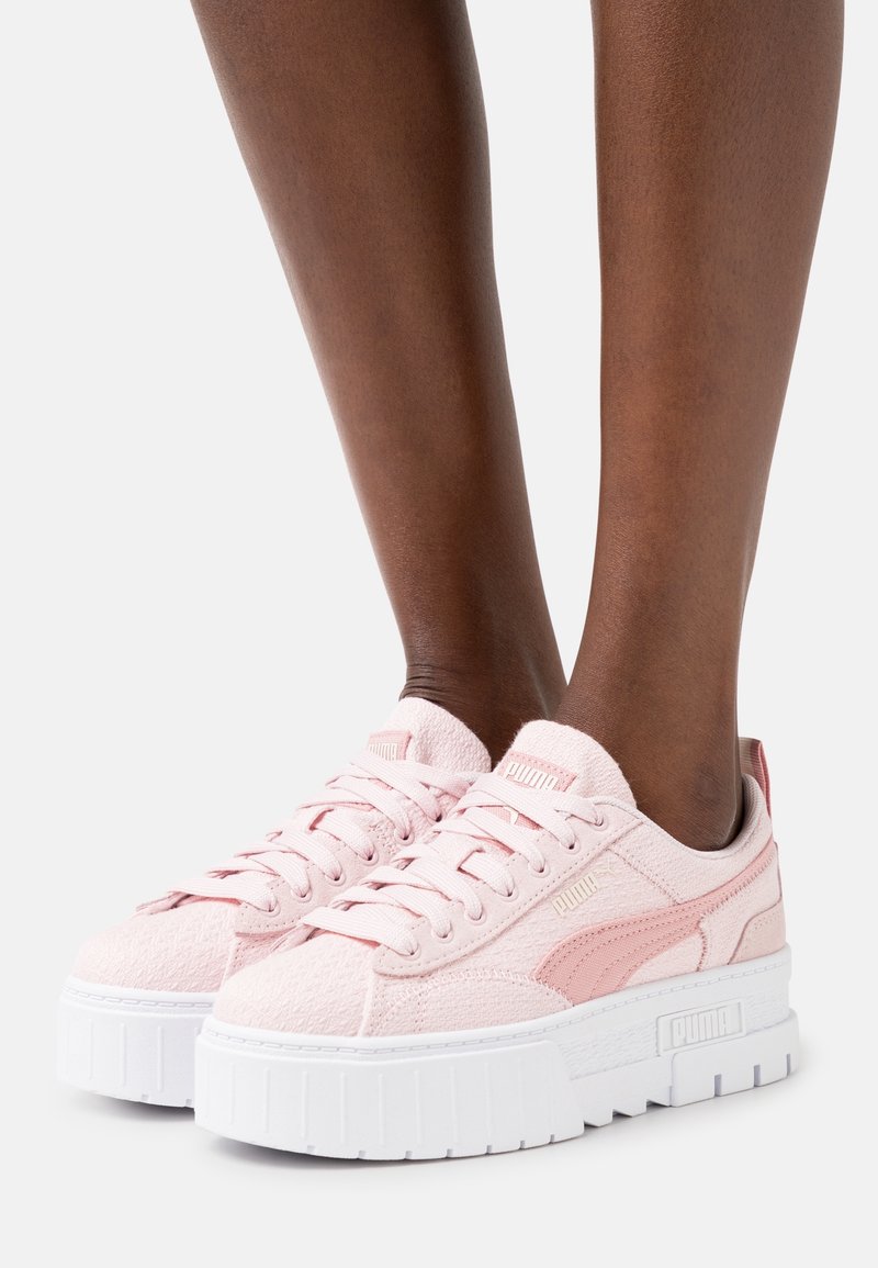 Jambes portant des baskets Puma rose clair avec des semelles plateformes épaisses blanches sur un fond clair uni.