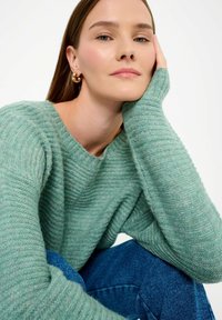 Maglione in maglia verde menta con scollo ampio e texture a coste, abbinato a jeans blu. Modella con il mento appoggiato sulla mano, mentre mostra il capo.