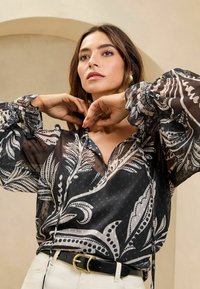 Bluză neagră transparentă cu un design floral în alb și bej, cu mâneci bufante, un șnur la decolteu și buline texturate.