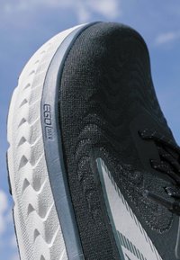 Scarpa sportiva nera con tomaia testurizzata, caratterizzata da una suola bianca a zigzag e accenti grigi. Presenta elementi di design moderni e branding.