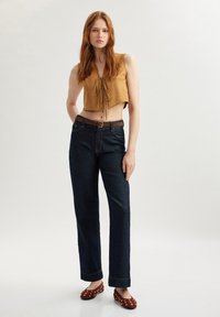 Haut en suédine taupe court avec un décolleté en V et un détail à nouer, associé à un jean droit bleu marine et des ballerines bordeaux avec des accents dorés.