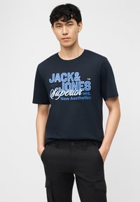 Giovane uomo con indosso una t-shirt grafica blu navy Jack & Jones e pantaloni cargo neri con le mani in tasca, in piedi su sfondo bianco.