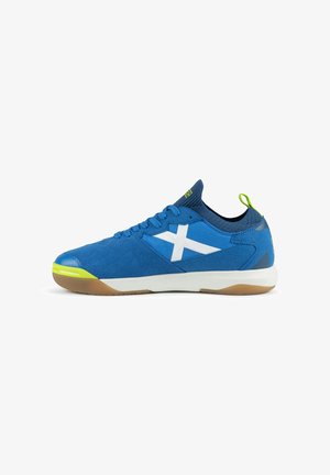 Zapatilla deportiva azul con cruz lateral blanca, detalles en la puntera y el talón en amarillo verdoso, cuello tipo calcetín azul oscuro y suela blanca con dibujo marrón.