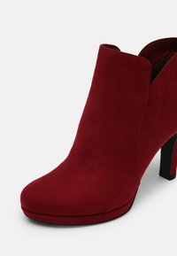 Bottine rouge en daim à talon haut avec bord supérieur incurvé et talon noir fin sur fond blanc.