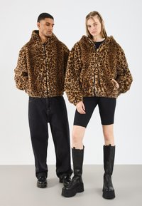 Două modele îmbrăcate în jachete de blană cu imprimeu de leopard maro; una cu pantaloni negri largi și încălțăminte, cealaltă cu pantaloni scurți negri și cizme cu platformă.