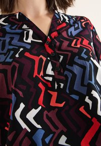 Bunt gemusterte Bluse mit einem abstrakten Muster in Rot, Blau, Weiß und Schwarz, mit einem V-Ausschnitt und einer Knopfleiste mit vier Knöpfen.