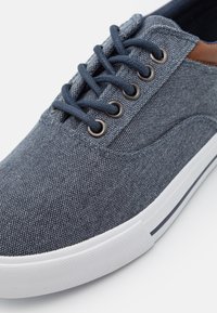Baskets en denim bleu avec une tige en tissu texturé, œillets en métal circulaires, lacets bleu marine, et une semelle en caoutchouc blanc avec une bande bleu marine.