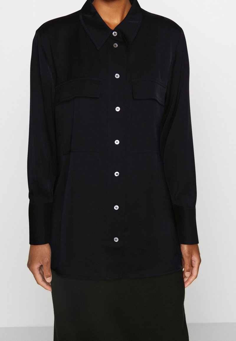 Camicia nera in satin con colletto classico, due tasche frontali, maniche lunghe e chiusura a bottoni. Texture liscia e vestibilità su misura.
