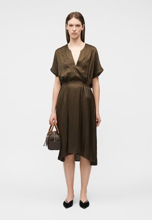 Žena v hnedých midi šatách s výstrihom do V drží malú hnedú kabelku s drevenými rukoväťami, doplnenú čiernymi balerínami s špicatou špičkou.