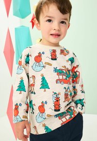 Chemise à manches longues de couleur beige clair avec des motifs de Noël colorés, y compris des arbres, des cadeaux et des personnages festifs, associée à un pantalon sombre.
