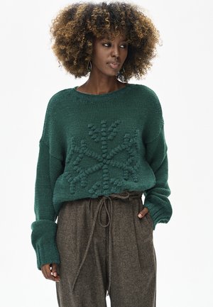 Grüner übergroßer Strickpullover mit strukturiertem Schneeflockenmuster, kombiniert mit braunen, hoch taillierten Hosen mit Bindegürtel.