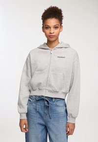 Donna con capelli ricci, indossa una felpa grigia con zip e tasche e jeans blu a vita alta con cintura, contro uno sfondo bianco semplice.
