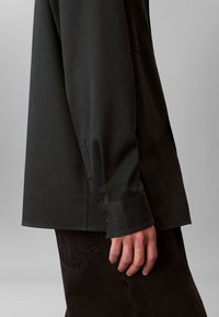 Camicia a maniche lunghe grigio scuro con vestibilità comoda, caratterizzata da uno spacco laterale e bottoni decorativi sul polsino. Abbinata a pantaloni marrone scuro con trama.