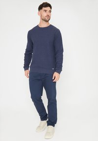 Sweter w kolorze granatowym, dzianinowy, z teksturowanym wzorem, okrągłym dekoltem i długimi rękawami, zestawiony z ciemnymi spodniami i jasnymi sneakersami.