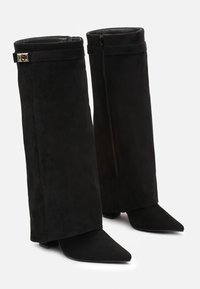 Bottes en daim noires à la cheville avec des bouts pointus, dotées d'une fermeture éclair et d'une lanière décorative avec un accent métallique en haut.