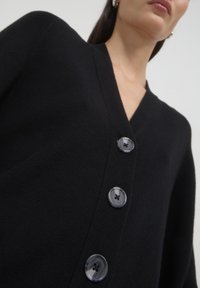 Schwarze Strickjacke mit V-Ausschnitt, die drei große graue Knöpfe hat. Der Stoff sieht weich und strukturiert aus und bietet eine zugleich strukturierte und entspannte Passform.