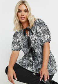 Top de estampado de piel de serpiente en blanco y negro con mangas abullonadas, tres lazos negros y un cuello redondo, combinado con pantalones negros.