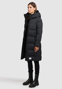 Zwarte puffer jas met capuchon, drukknoopsluiting, zijzakken en een doorgestikte textuur. Gemaakt over zwarte leggings en enkellaarzen.