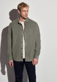 Camisa de botones verde claro con un bolsillo en el pecho, cuello desabrochado y mangas largas. Lleva debajo una camiseta beige y se combina con pantalones oscuros.