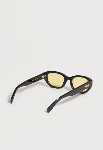 Lunettes de soleil rectangulaires noires avec verres teintés jaunes et « Angels » en jaune sur la branche, vues de dos sur une surface blanche.