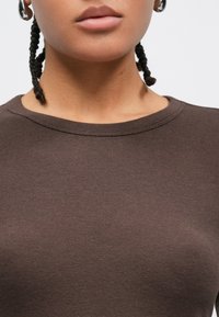 Καφέ βαμβακερό μπλουζάκι με στρογγυλή λαιμόκοψη, με λεία υφή. Το ύφασμα φαίνεται απαλό και εφαρμοστό στο σώμα. Χωρίς εμφανή σχέδια.